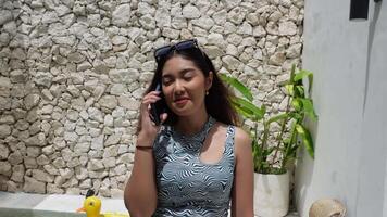 Indonesian woman wearing a pattern sleeveless top smiling while talking on phone call and walking at villa, working on holiday video