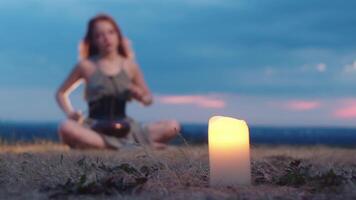 Red-haired girl is playing the glucophone, out of focus, in nature, sitting on the grass, against the backdrop of a burning candle. Full shot. Slow motion. video