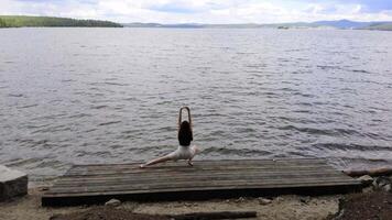 un' giovane ragazza è praticante gli sport e allungamento su un' di legno piattaforma, collocato su il riva di un' grande lago, a il piede di montagne circondato di pino alberi, su un' nuvoloso giorno. aereo filmato. 4k video