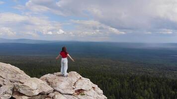 Jeune fille des stands sur Haut de une Montagne dans le pluie, dans de face de une énorme forêt à le horizon, elle se répand sa bras en dehors dans le été pluie, profiter le cool brise et rafraîchissant pluie. aérien images. video