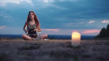 Beautiful red-haired girl, dressed in a green gown, plays a steel tongue drum in nature against a backdrop of a stormy sky, sitting on the grass under the soft glow of candlelight. Full shot. Slow motion. 4k video