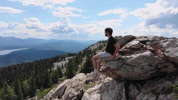 A man is enjoying a beautiful view of the mountains. He is sitting on a hilltop, taking in the scenery. The air is crisp and fresh, and he can smell the scent of pine trees. Aerial footage. 4k video