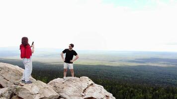 Man and a woman take pictures of each other, standing on top of a mountain with the forest, river, and Ural Mountains in the background. Aerial footage. 4k video