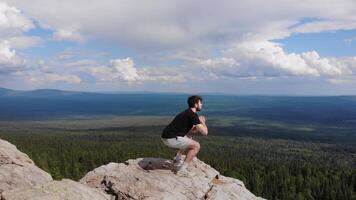 uomo è fare squat e esercizio su il bordo di un' scogliera nel il montagne, con un' foresta come un' sfondo. aereo filmato. 4k video