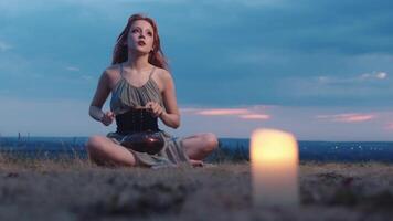 Beautiful red-haired girl in a green dress is playing a tongue drum in nature, sitting on the grass under the candlelight. She seems to be enjoying the music she is playing. Full shot. Slow motion. video