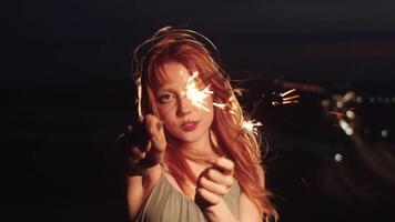 Beautiful red-haired woman with bright makeup looks into the camera, holding a sparkler as she stands against the backdrop of the blurred city lights. Medium shot. Slow motion. video