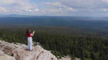 A beautiful girl, wearing a red T-shirt, photographs a forest during the summer rain, from the top of the mountain. She is standing on the edge of the cliff on a cloudy day. Shooting from a drone. 4k video