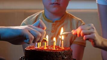Little birthday boy, wearing a birthday cone hat, holds a chocolate cake in his hands. The candles on the cake are lit by his loving parents, and he smiles with joy. Mid shot. 4k video
