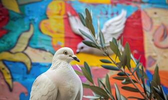 Close-up of a white pigeon with its wings folded in front of a colorful mural depicting a dove carrying an olive branch as a symbol of peace photo