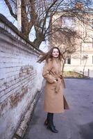 a girl in a brown coat is happy  near a white wall on a spring day photo