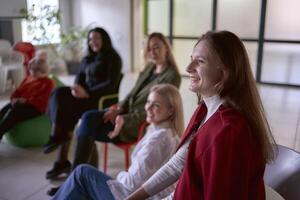 a woman makes a speech on stage, colleagues laugh at her jokes sitting on chairs and ottomans in the hall, office location photo