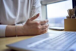 middle age woman with blond hair drinks coffee and works at a laptop in a coworking space with panoramic windows and a view of the city from above, details laptop, coffee, table photo