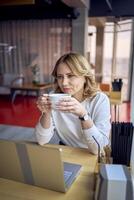 young blonde woman drinking coffee and working on a laptop in a cafe with panoramic windows and a view of the city from above photo