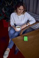 beautiful middle-aged woman drinking coffee in a beautiful cafe with greenery and light panels in the background, a phone with a green screen on the table photo