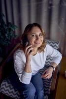 a woman in a sweater and jeans is talking on the phone in a beautiful cafe with greenery and light panels in the background photo