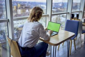 middle age woman working on a laptop in a coworking space with panoramic windows and a view of the city from above, laptop with a green screen, Chroma key photo