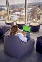 a woman in casual clothes is sitting at a laptop on an ottoman in front of a panoramic window with a view of the city, green screen, Chroma key photo