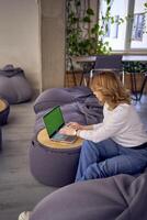 a woman in casual clothes is sitting at a laptop on bean bag in front of a panoramic window with a view of the city, green screen, Chroma key photo