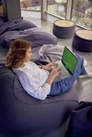 a woman in casual clothes is sitting at a laptop on an ottoman in front of a panoramic window with a view of the city, green screen, Chroma key photo