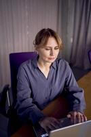 hermosa medio años concentrado negocio mujer vistiendo gris camisa y utilizando ordenador portátil mientras sentado en moderno espacio de trabajo foto