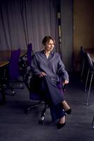 beautiful middle age business woman wearing gray shirt, wide leg pants abd black stilettos sit in purple office chair in modern workspace photo