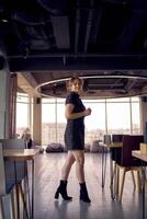 atractivo mujer en un negro mini vestir negocio estilo camina en un moderno trabajo colaborativo espacio con panorámico ventanas y un negro techo foto