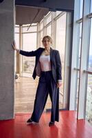 atractivo medio años mujer en chaqueta y blanco camiseta posando en moderno oficina con panorámico ventanas foto