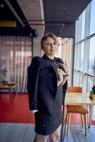 attractive woman in a black business style mini dress throws a jacket over her shoulders in a modern coworking space with panoramic windows and a black ceiling photo