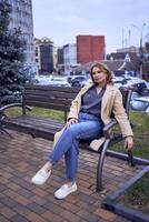 joven adulto mujer en pantalones y arena Saco sentado en un banco en un primavera día, moderno arquitectura detrás foto