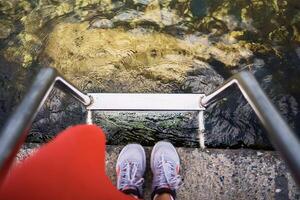 niña en un rojo vestir y zapatillas en el muelle mira a el claro agua foto