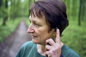 elderly woman engaged in Nordic walking with sticks in spring forest switches music on headphones photo