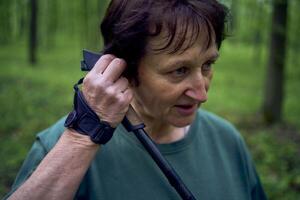 elderly woman engaged in Nordic walking with sticks in spring forest switches music on headphones photo