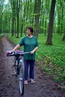 mayor mujer caminando en primavera bosque con bicicleta y yoga estera foto