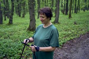 elderly woman doing Nordic walking with sticks in spring forest switches podcast on phone photo