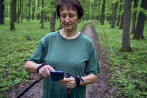 elderly woman doing Nordic walking with sticks in spring forest switches podcast on phone photo