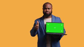 Salesman holding isolated screen laptop, doing recommendation, talking with audience. BIPOC person pointing towards chroma key notebook and showing thumbs up sign, studio background, camera B video