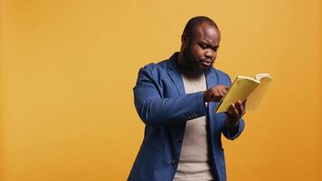 Man struggling to read book with small font, pointing finger to words on page. Person enjoying literature novel, wetting fingers to change page, isolated over studio background, camera B video