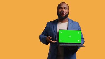 Smiling man holding isolated screen laptop, doing recommendation, talking with viewers. Cheerful BIPOC person pointing towards chroma key notebook, studio background, camera B video