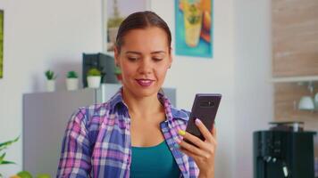 caucasico allegro donna scorrimento su smartphone e potabile verde tè nel il mattina a prima colazione. Tenere Telefono dispositivo con touch screen utilizzando Internet tecnologia navigando, ricerca su gadget. video