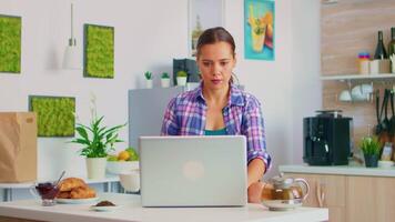 de bonne humeur Dame sirotant vert thé et envoi postal sur portable ordinateur pendant petit déjeuner dans confortable cuisine. travail de Accueil en utilisant dispositif avec l'Internet technologie, navigation, recherche sur gadget dans le Matin. video