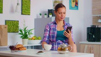 donna navigazione su smartphone mentre potabile verde Tè, nel il mattina durante prima colazione. Tenere Telefono dispositivo con touch screen utilizzando Internet tecnologia scorrimento, ricerca su intelligente gadget. video