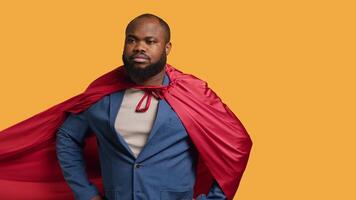 African american superhero flying with red cape, isolated over studio background, flexing muscles. Man wearing cloak posing as hero in costume showing courage and strength, camera A video