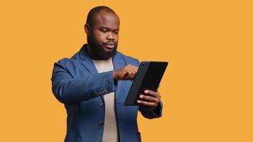 Man happily scrolling on tablet touchscreen to check social media feed. African american person browsing internet websites, entertained by content, studio background, camera A video