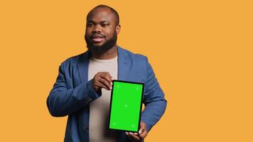 Amused african american man laughing while presenting tablet with green screen display, studio background. Joyous BIPOC person has fun during promotion for blank copy space mockup device, camera A video