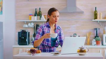 donna sorseggiando verde tè e digitando su sua il computer portatile computer durante prima colazione nel accogliente cucina. Lavorando a partire dal casa utilizzando dispositivo con Internet tecnologia, navigando, ricerca su aggeggio nel il mattina. video