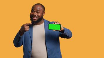 Portrait of man doing influencer marketing using isolated screen phone, showing thumbs up sign, studio background. Merry BIPOC person holding empty copy space mockup cellphone, camera A video