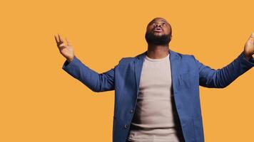 Cheerful pious african american man looking up to sky, waiting for sign from his god. Happy spiritual man doing worship hand gesturing, hoping for miracle, isolated over studio background, camera B video