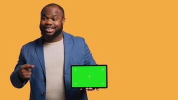 Upbeat african american man holding green screen tablet, doing recommendation. Cheerful BIPOC person pointing towards mockup device, giving positive feedback, studio background, camera A video