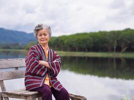 retrato de un mayor mujer sonriente, brazos cruzado y mirando a el cámara mientras sentado lado el lago. espacio para texto. concepto de Envejecido personas y relajación foto