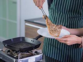 Close-up of cooking a steak by an Asian young man and a young woman wearing an apron and making a steak in the kitchen in his home, grill the steak on a small gas stove in the kitchen. Food concept photo